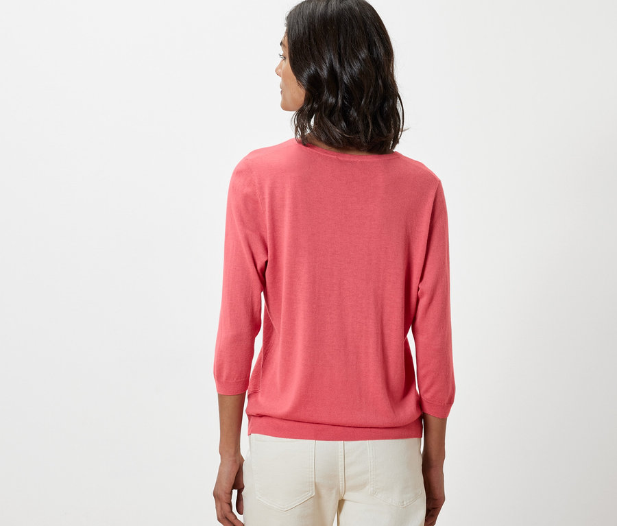 Žena má na sobě korálový svetr z jemného úpletu s hedvábím a bílé kalhoty.