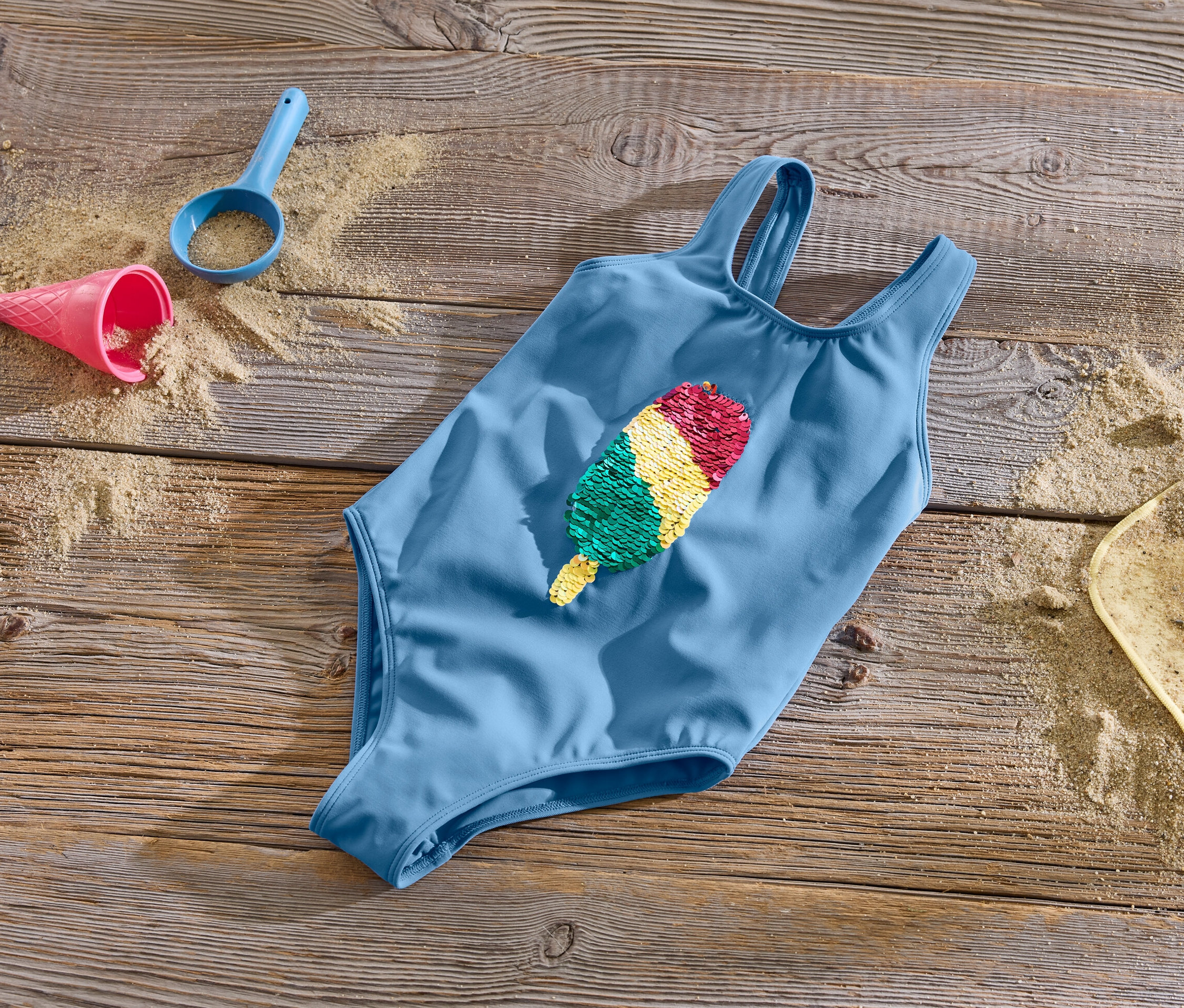 Modré dívčí plavky s oboustrannými flitry leží na dřevěném povrchu s pískem a hračkami.