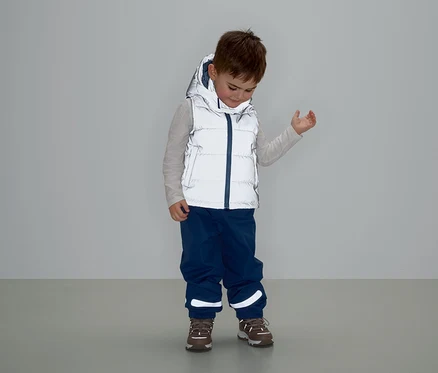 Chlapec v šedé vestě s kapucí, modrých kalhotách a hnědých botách stojí a dívá se dolů.