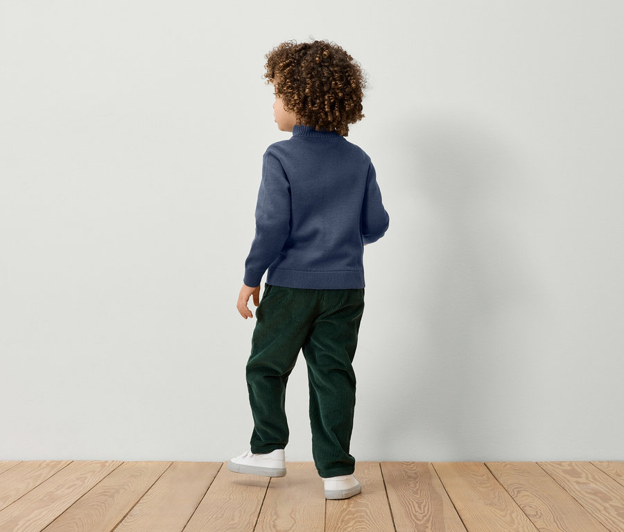 Chlapec s kudrnatými vlasy stojí zády a má na sobě modrý svetr, zelené kalhoty a bílé tenisky.