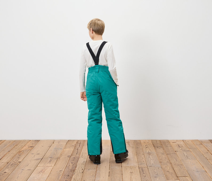 Chlapec stojí zády k fotoaparátu a má na sobě tyrkysové lyžařské kalhoty s černými šlemi.