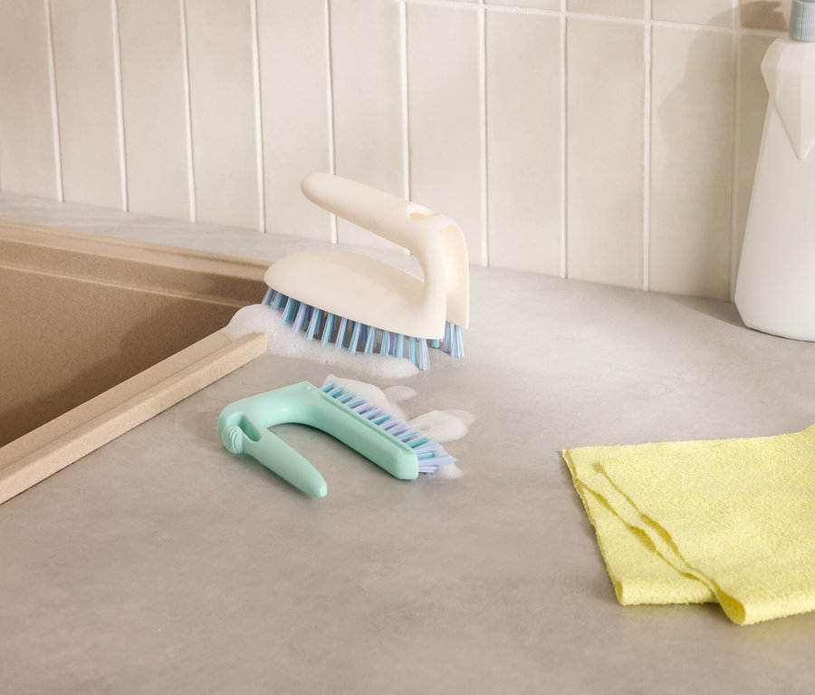 Dva kartáče na čištění a žlutý hadřík na šedé desce stolu vedle dřezu.