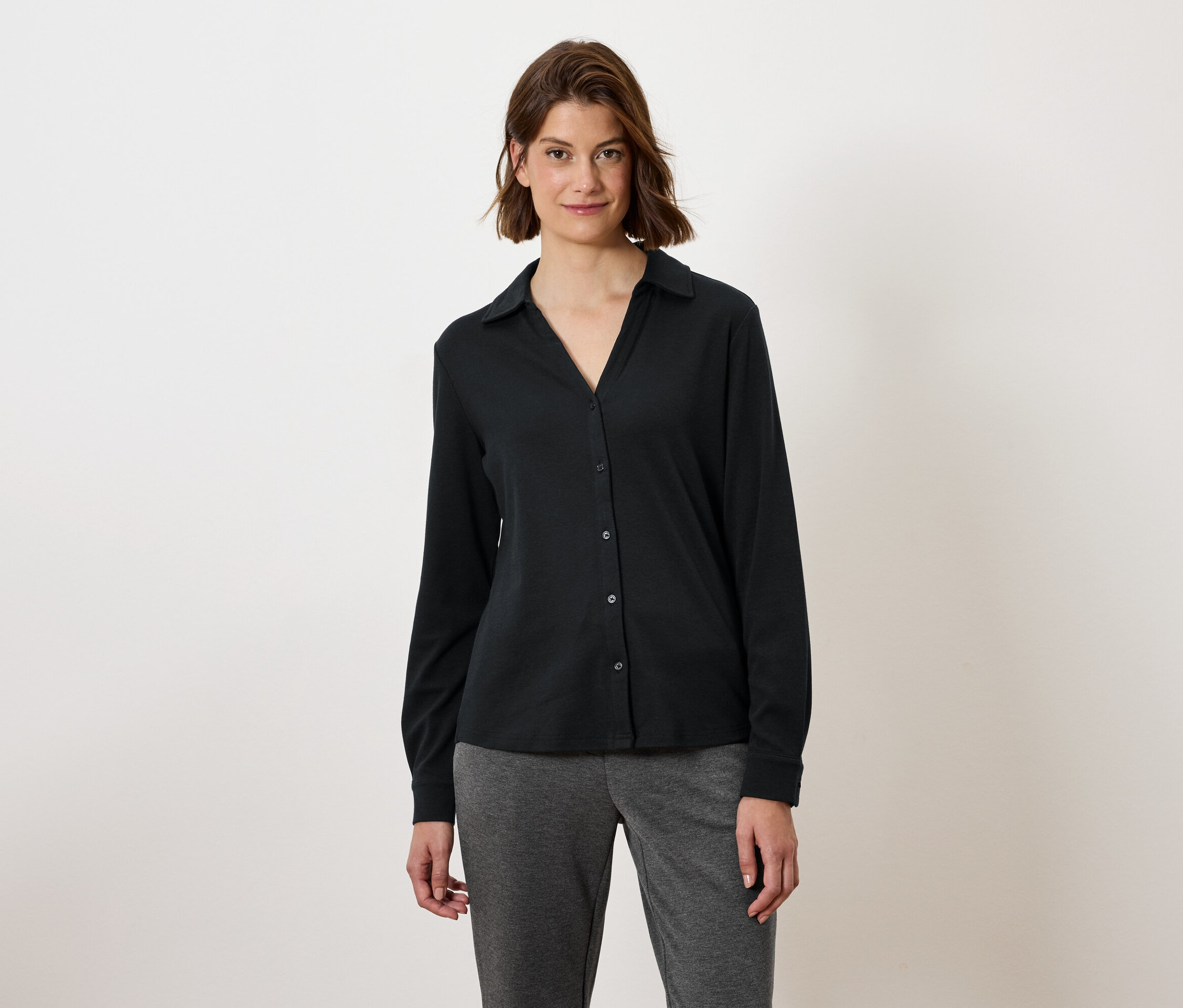 Žena s hnědými vlasy nosí černou košili s dlouhým rukávem a šedé kalhoty.