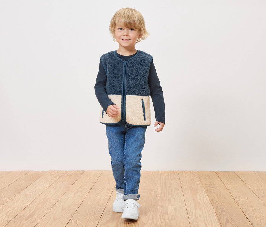 Chlapec v modré fleecové vestě s krémovými spodními panely na zip stojí na dřevěné podlaze.