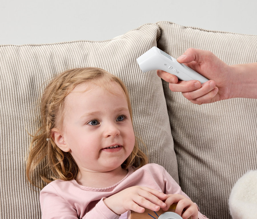 Malá holčička leží na pohovce, zatímco ruka drží bílý digitální teploměr u jejího čela.