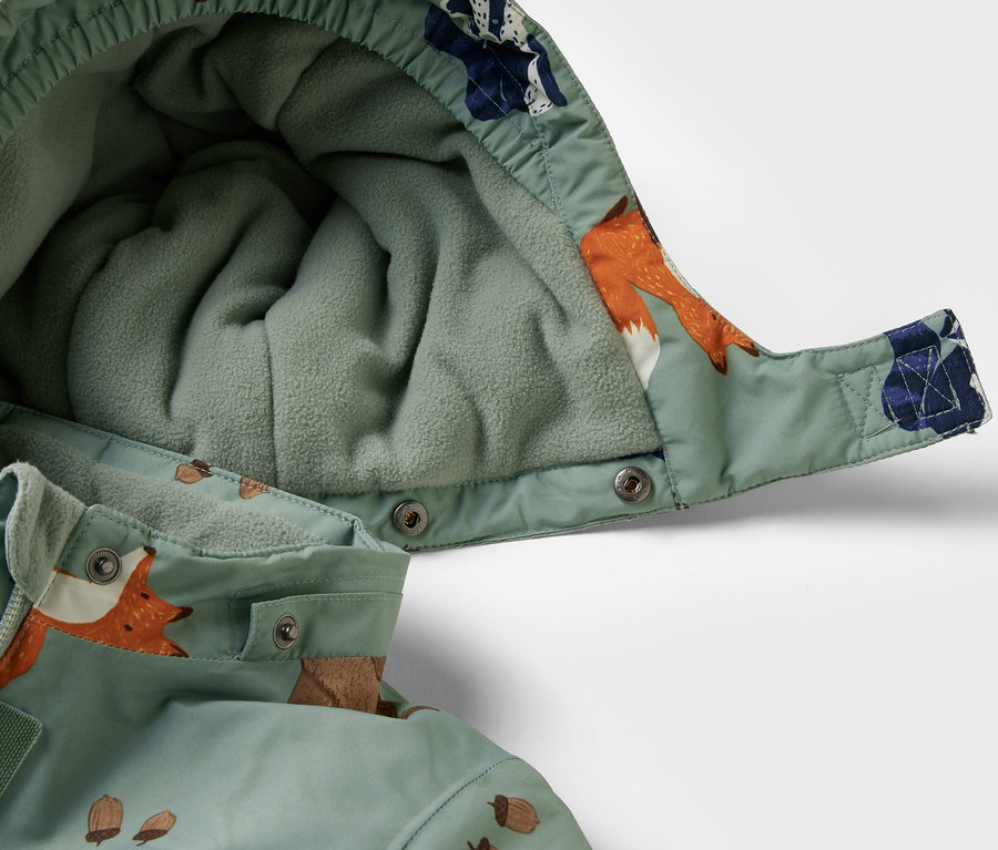 Detail dětské zimní kombinézy s kapucí a vzorem lišek a žaludů.
