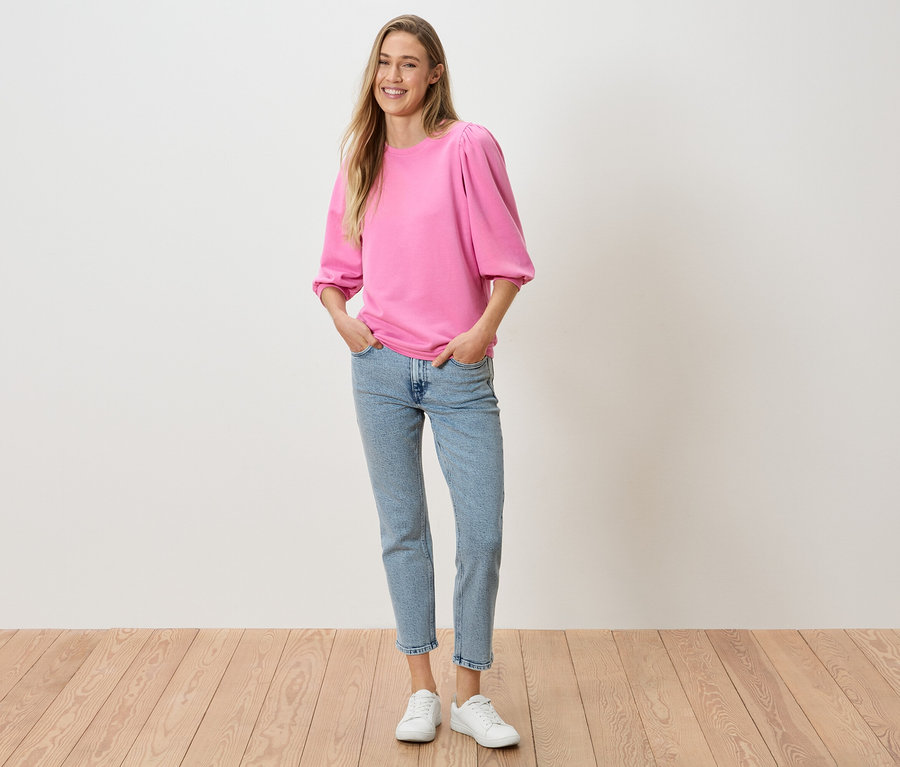 Žena s rukama v kapsách kalhot má na sobě růžový top s nabíranými rukávy, džíny a bílé tenisky.