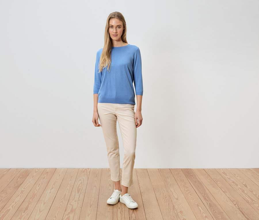 Žena stojí před bílou zdí, má na sobě modrý svetr, světlé kalhoty a bílé boty.