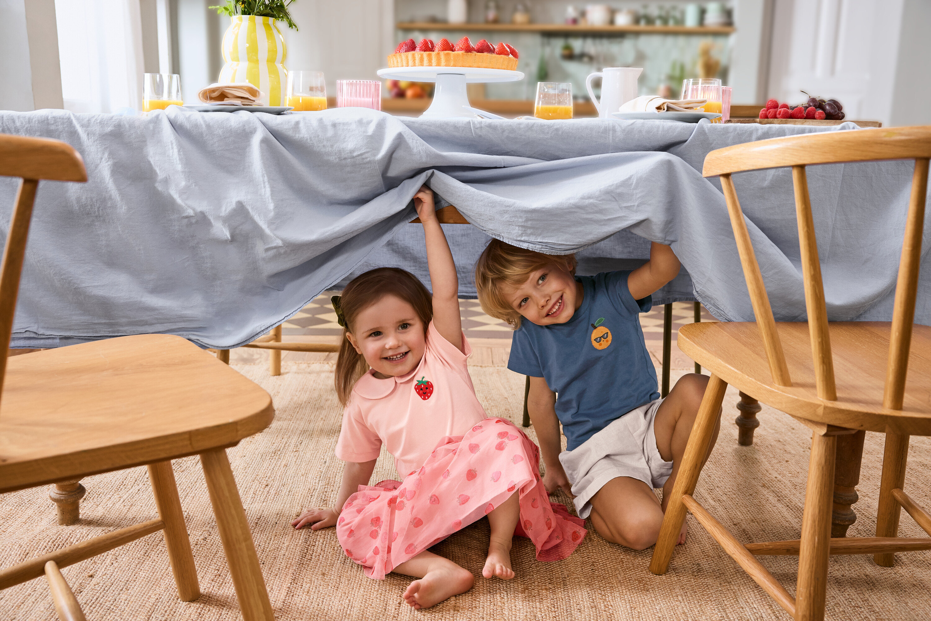 Dvě děti sedí pod stolem se světle modrým ubrusem. Dívka má na sobě Dětskou tylovou sukni a 2 Dětská trička. Chlapec má na sobě Dětské šortky.