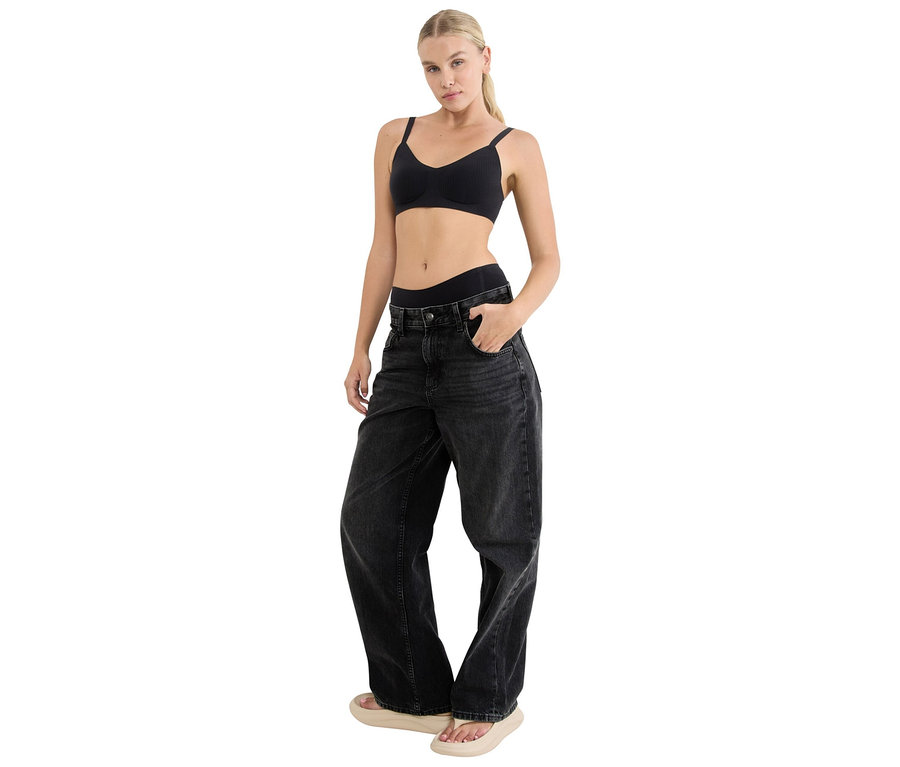 Žena v černé měkké podprsence sloggi ZERO Feel Pure Soft bra a černých širokých džínách, stojí s rukou v kapse.