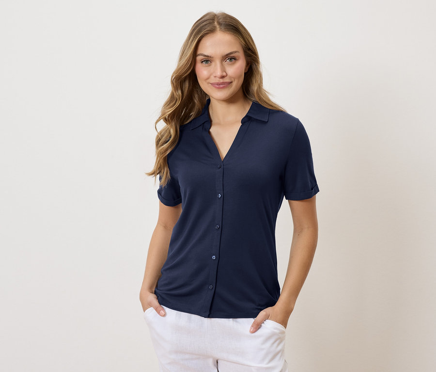 Žena s dlouhými vlnitými blond vlasy v tmavě modré košili a bílých kalhotách má ruce v kapsách.