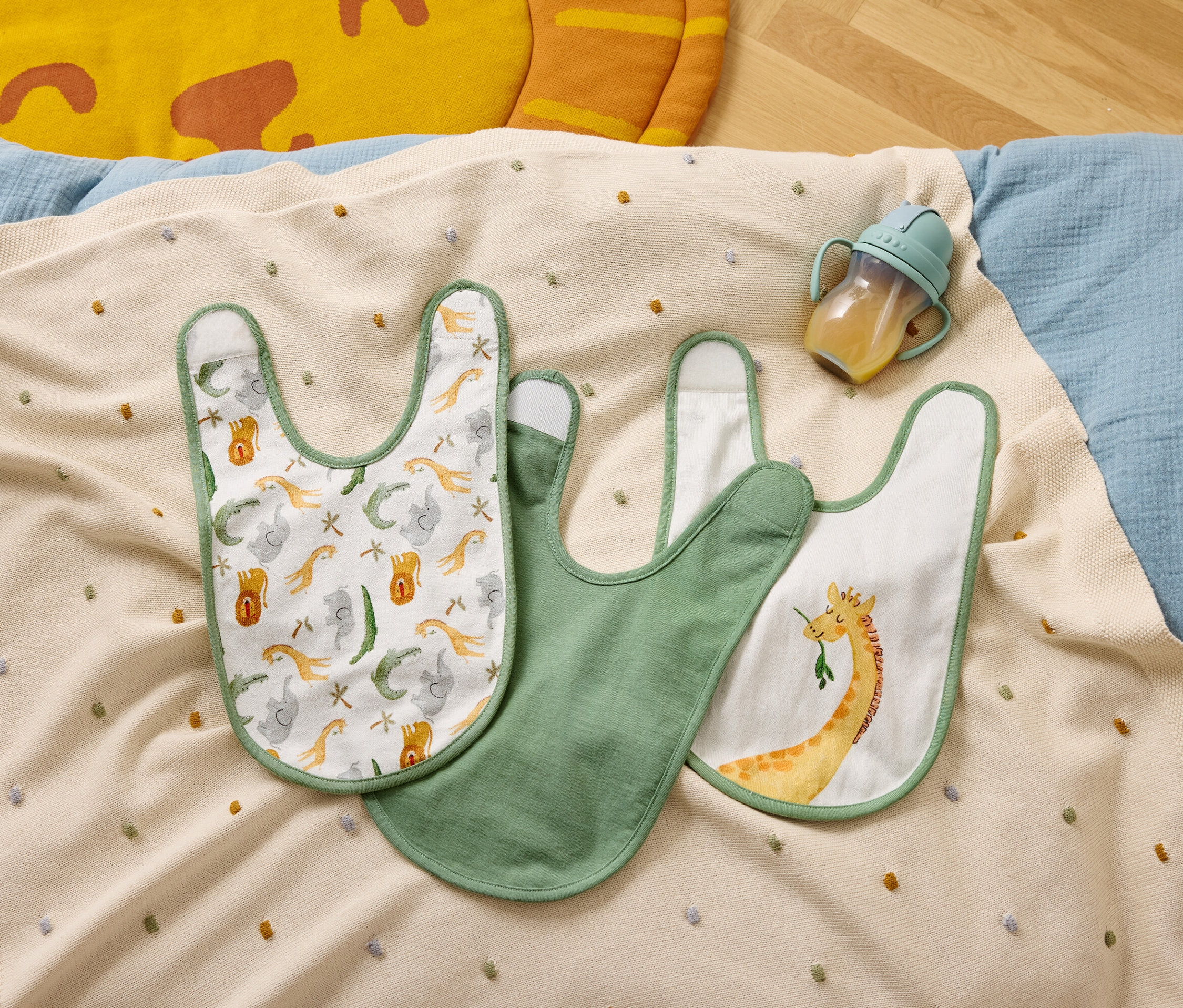 Tři bryndáčky a láhev na pití leží na béžové dece s barevnými puntíky.