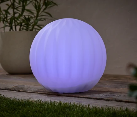 Kulatá, fialová lampa stojí na dřevěném stole vedle hrnkové rostliny.