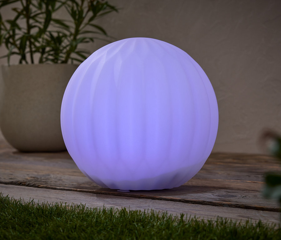 Kulatá, fialová lampa stojí na dřevěném stole vedle hrnkové rostliny.