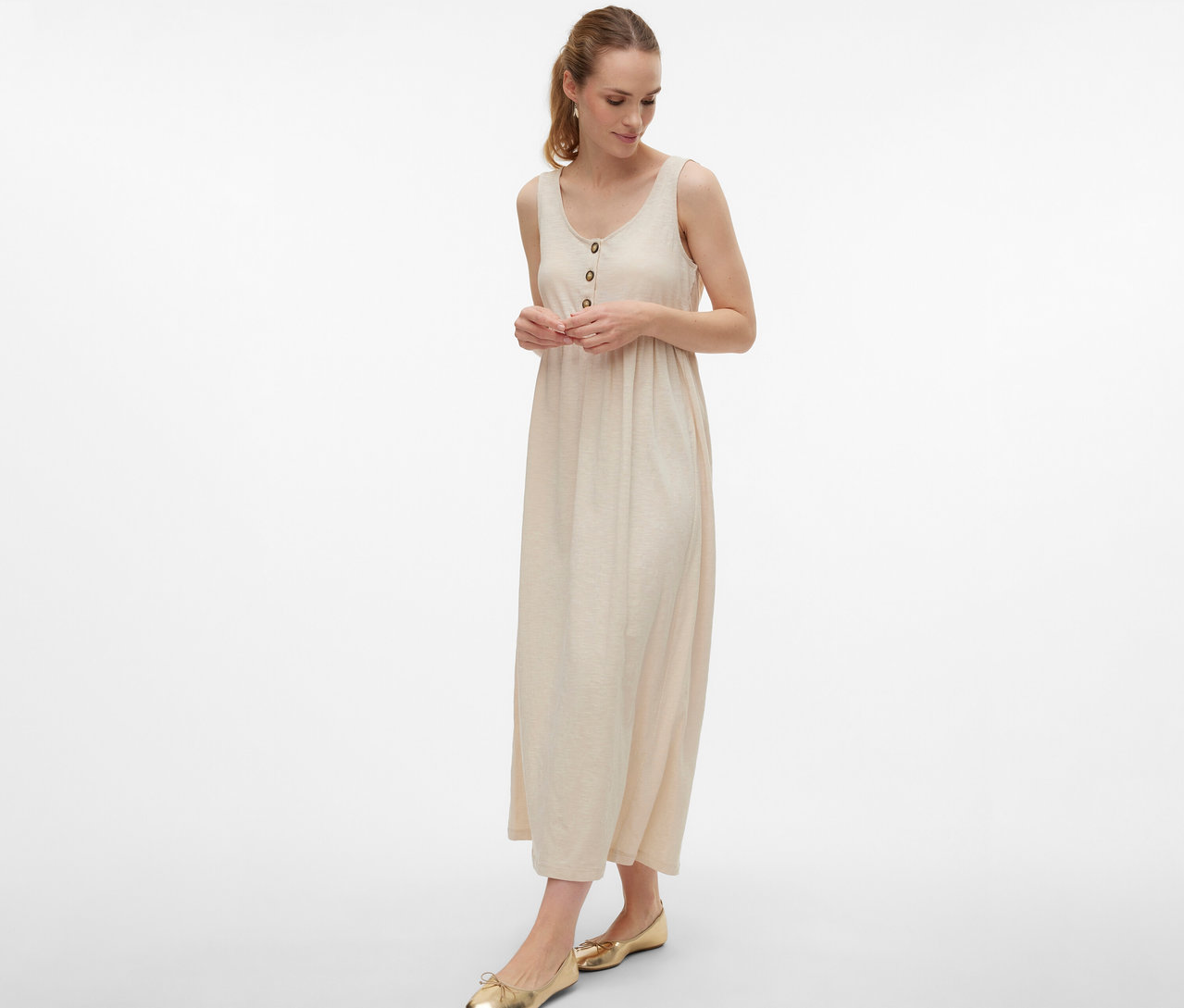 Žena nosí těhotenské šaty MAMALICIOUS »MLEVI LIA« s knoflíky a zlaté baleríny na bílém pozadí.