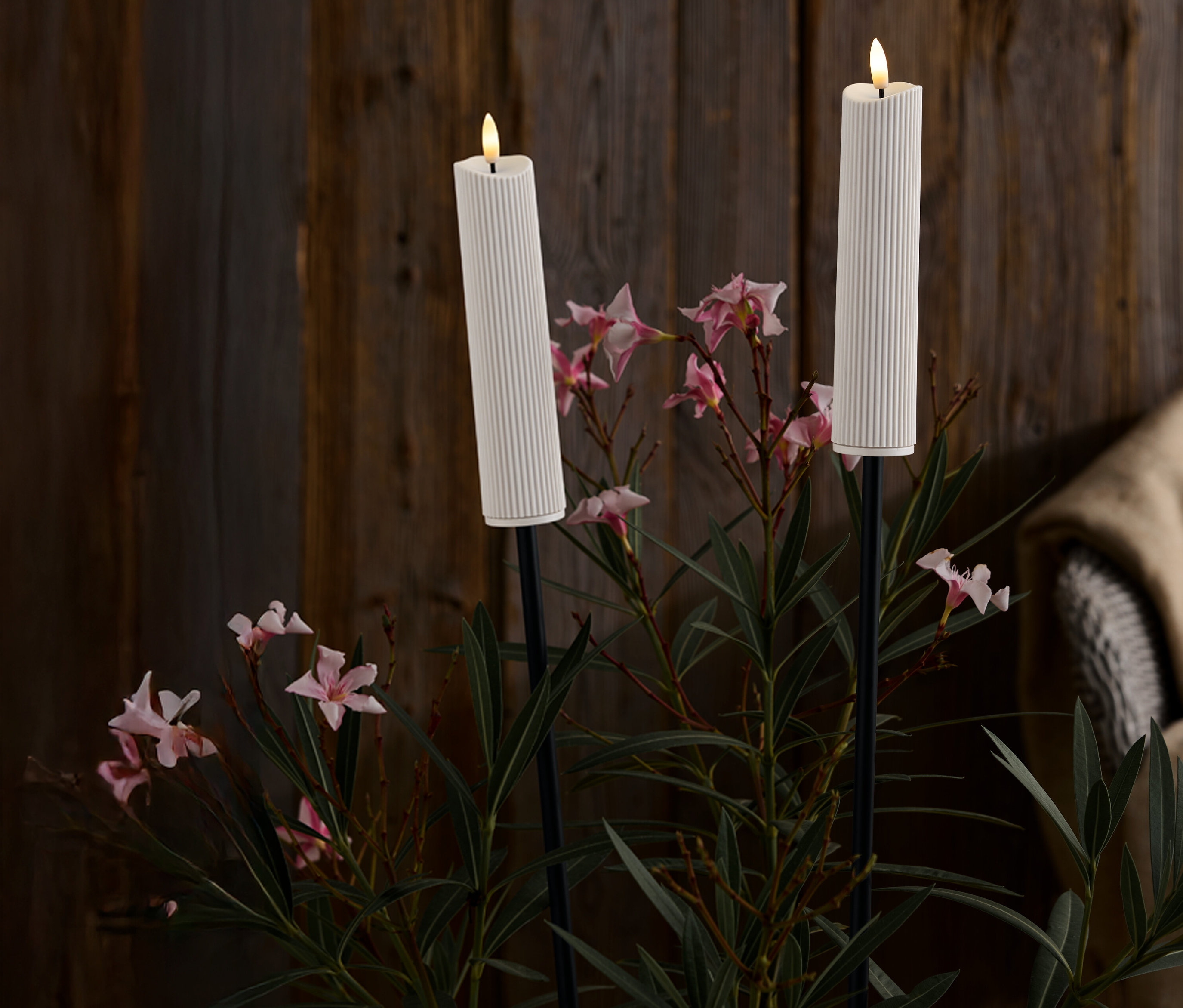 Dvě bílé, žebrované svíčky na černých stojanech uprostřed růžových květů a zelených listů na pozadí tmavého dřeva.