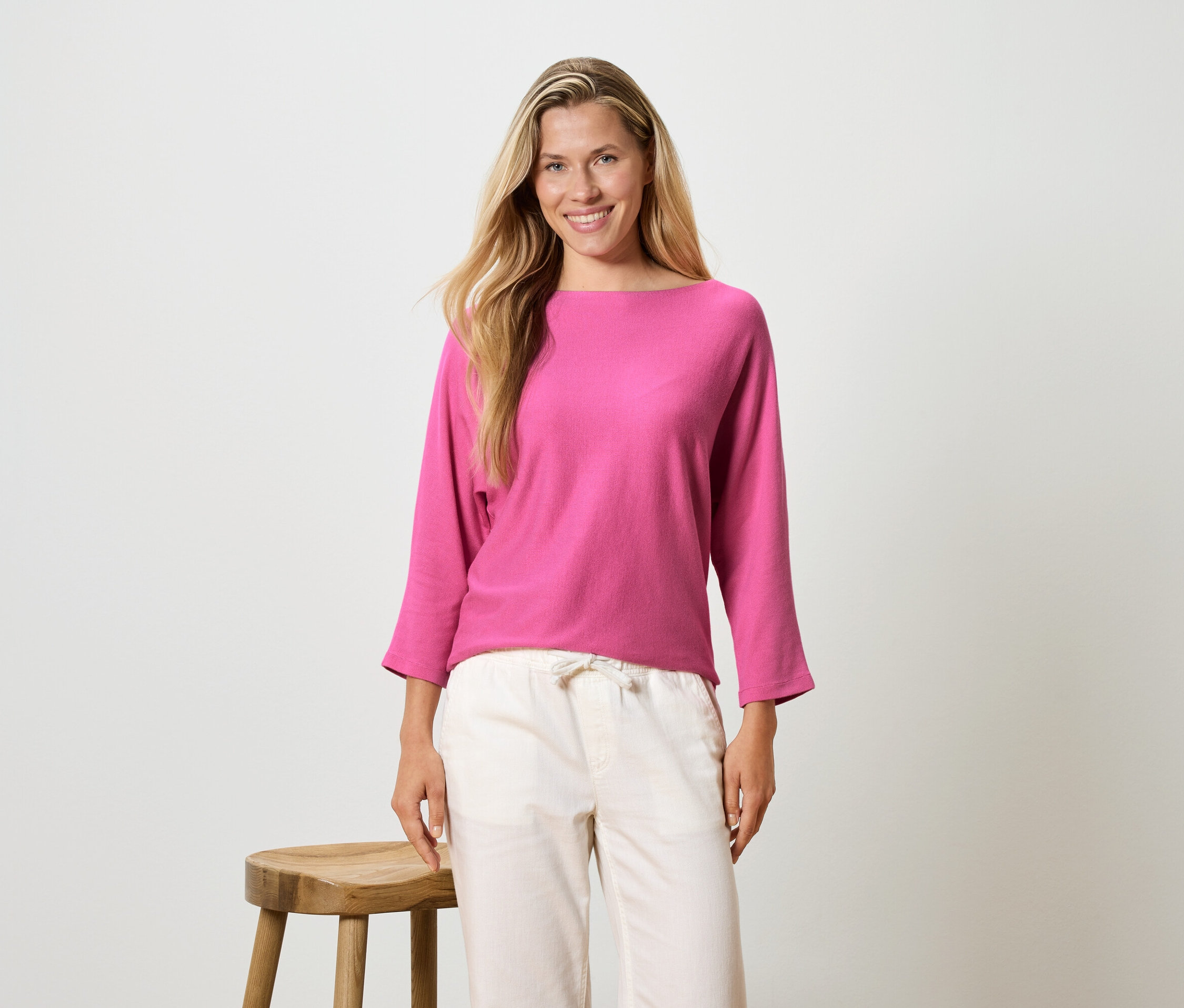 Žena s dlouhými blond vlasy má na sobě růžovou blůzu s dlouhým rukávem a světlé kalhoty, stojí vedle stoličky.