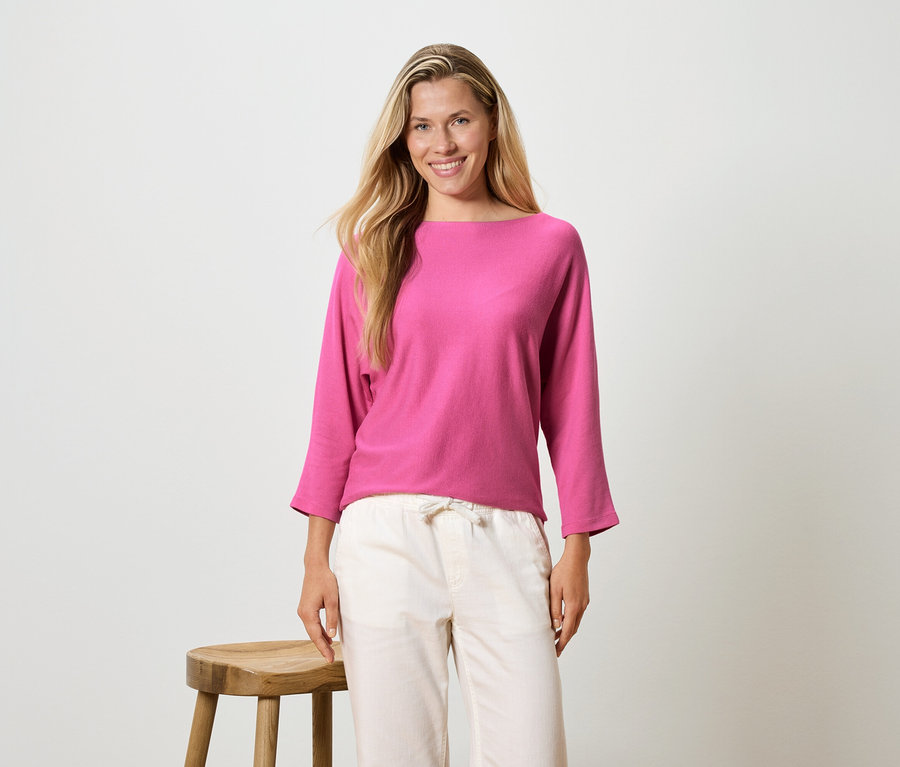 Žena s dlouhými blond vlasy má na sobě růžovou blůzu s dlouhým rukávem a světlé kalhoty, stojí vedle stoličky.