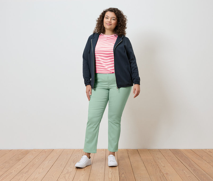 Žena stojí ve světlém prostředí a má na sobě tmavě modrou bundu, pruhovaný top, světle zelené kalhoty a bílé tenisky.