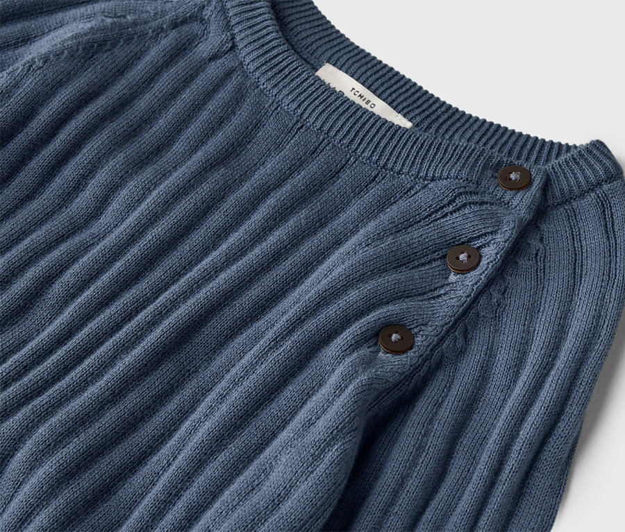 Detailní záběr na žebrovaný, tmavě modrý pletený svetr se třemi knoflíky u krku.