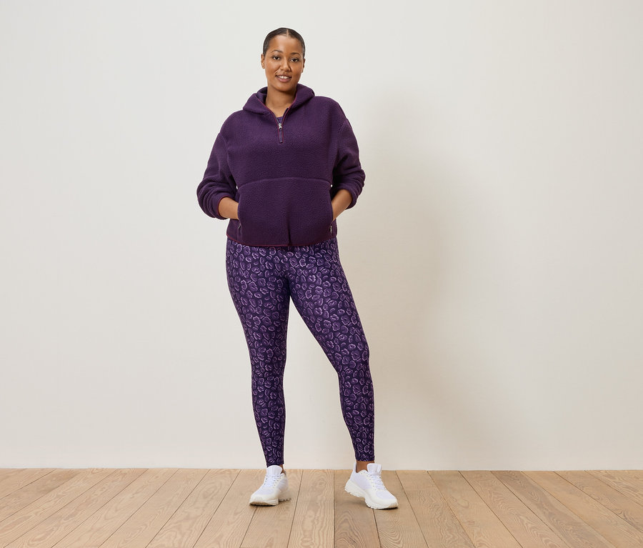 Žena v purpurové fleecové mikině na poloviční zip a purpurové legíny s leopardím vzorem, která má na sobě bílé tenisky.