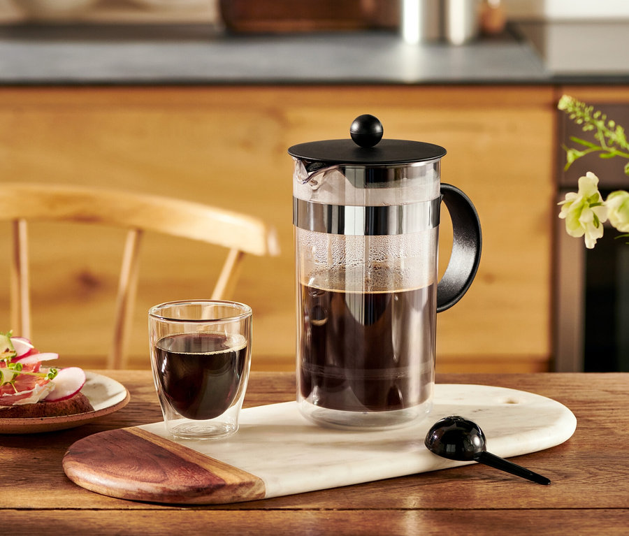 Kávovar french press Bodum® 1 l stojí na stole vedle šálku kávy a talíře s chlebíčky.