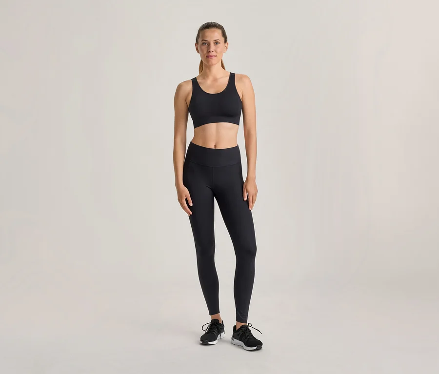 Žena stojí v černé sportovní podprsence, černých legínách a černých sportovních botách před světlým pozadím.