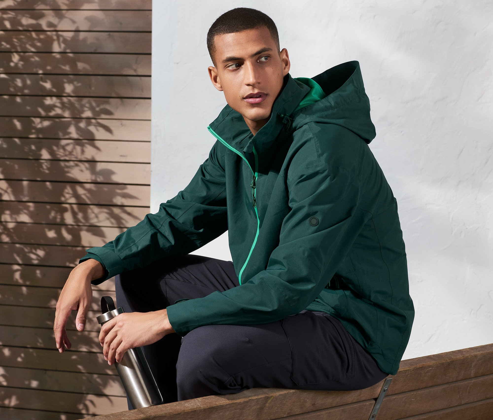 Muž sedí na lavičce a drží kovovou láhev. Má na sobě outdoorovou bundu.