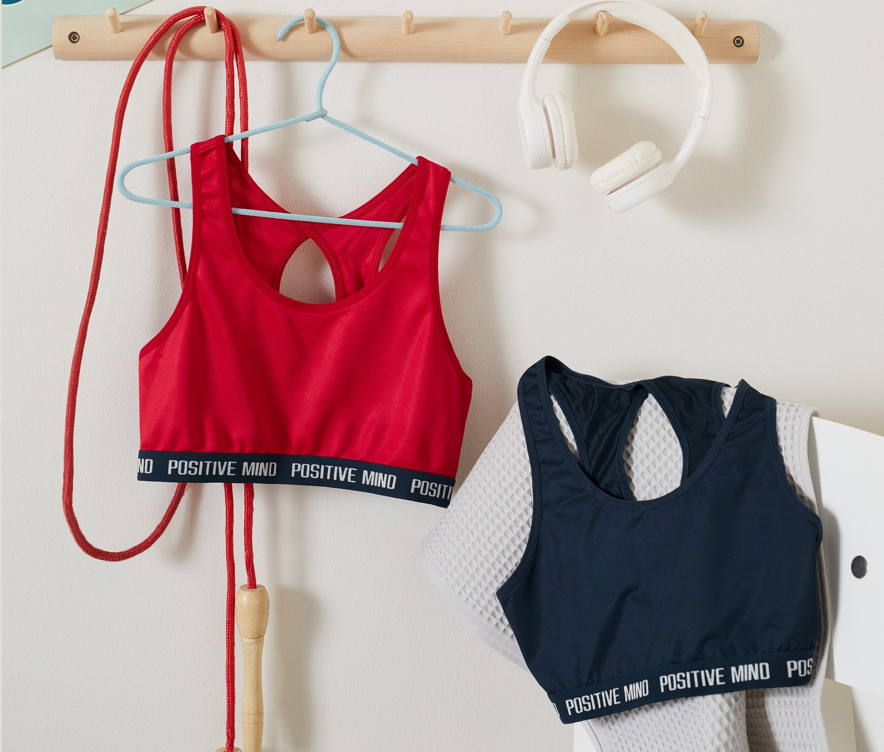 Červená sportovní podprsenka z 2 ks visí na ramínku vedle švihadla. Modrá sportovní podprsenka z 2 ks leží na šedé židli vedle ručníku.