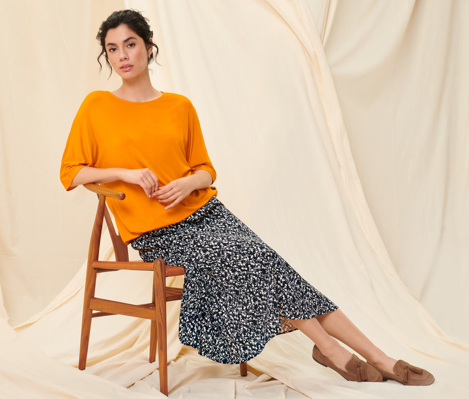 Žena sedí na dřevěné židli, má na sobě oranžové halenkové triko, květovanou midi sukni a boty z velurové kůže.