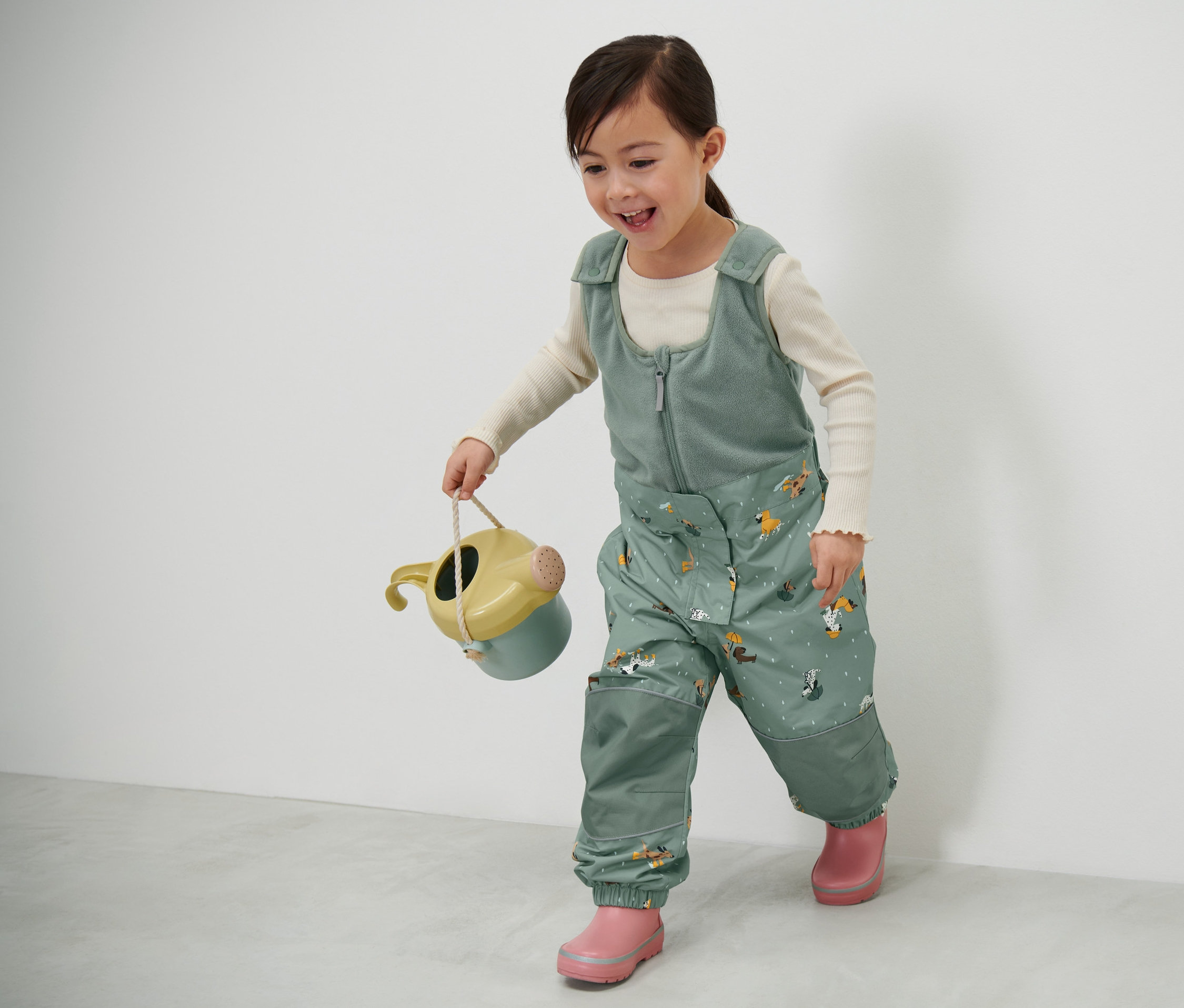 Holčička nese konev a má na sobě dětské termo kalhoty do deště.