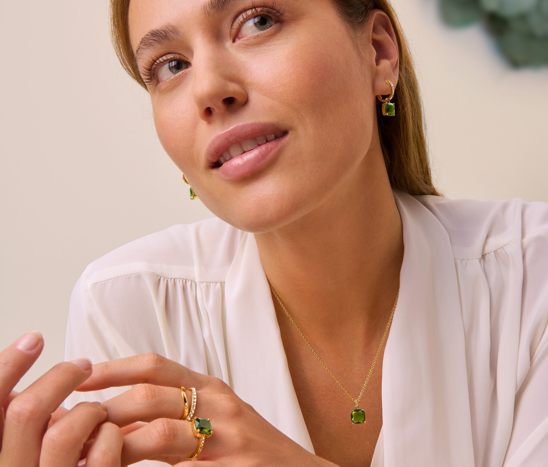 Žena má na sobě náhrdelník Forest se zeleným skleněným kamínkem, kreolky Forest se zeleným skleněným kamínkem a prsten Forest se zeleným skleněným kamínkem a prsten s dvojitou vlnou a kamínky.