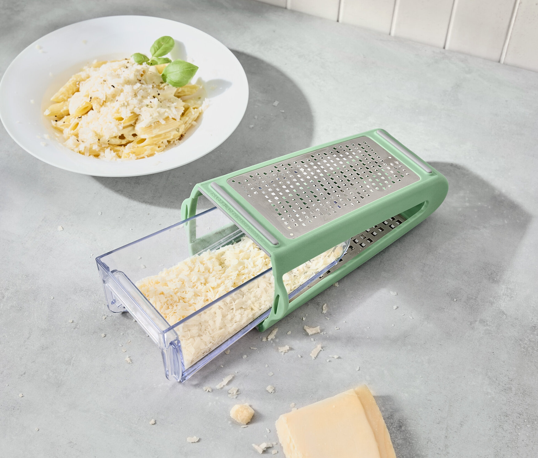 Sýr se strouhá pomocí obousměrného struhadla se záchytnou nádobkou. V pozadí je talíř těstovin.