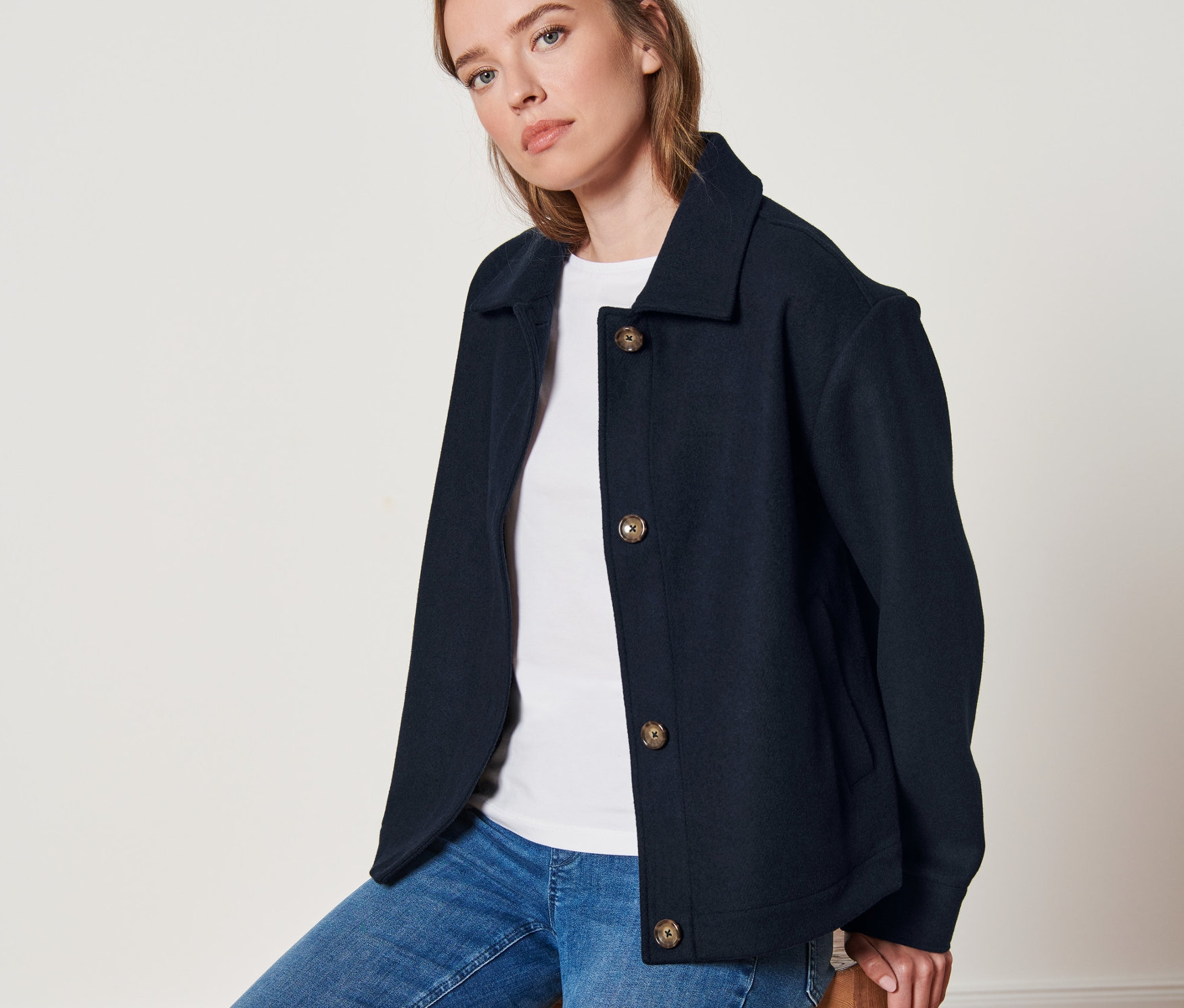 Žena sedí na dřevěné stoličce a má na sobě krátkou bundu na přechodné období.