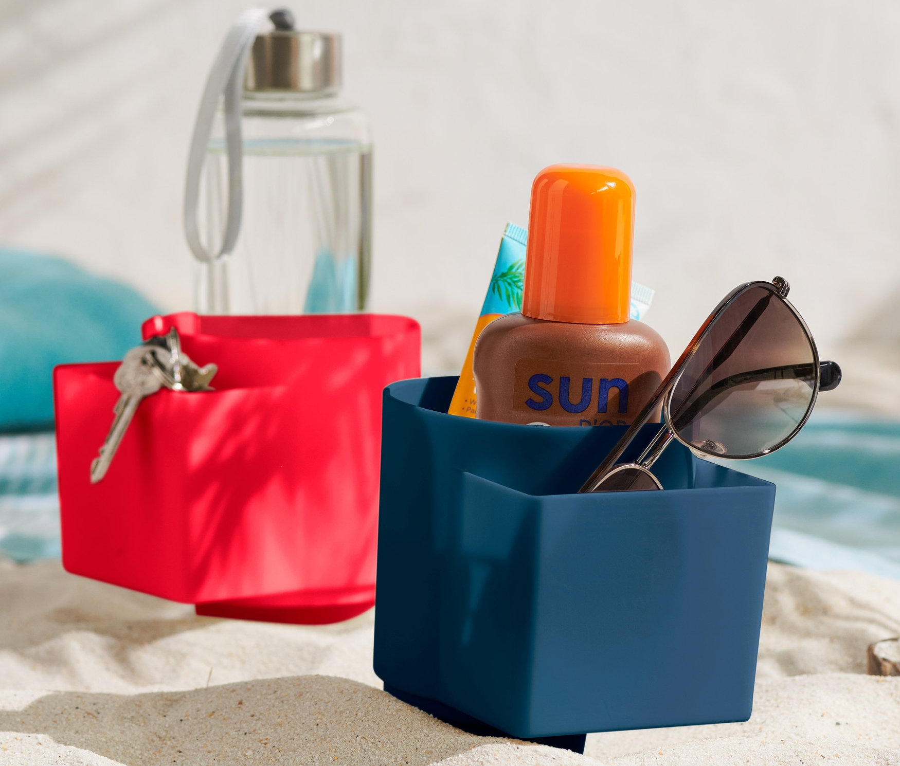 Dva plážové držáky na pití na písku. V modrém držáku jsou sluneční brýle a opalovací krém.