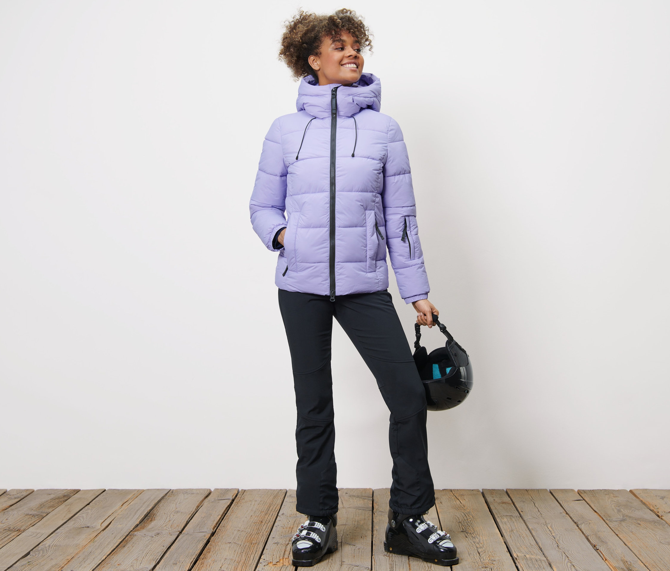 Žena v prošívané lyžařské bundě, černých kalhotách a lyžařských botách drží helmu.