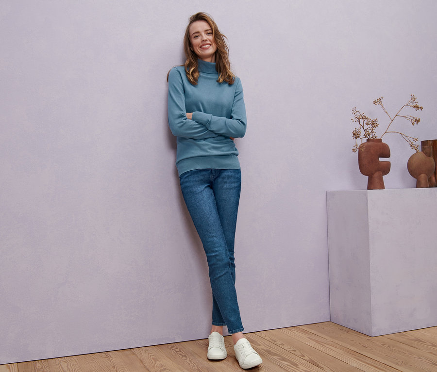 Žena pózuje v kouřově modrém roláku z jemného úpletu, skinny džínách »Fit Hanna« a kožených teniskách.