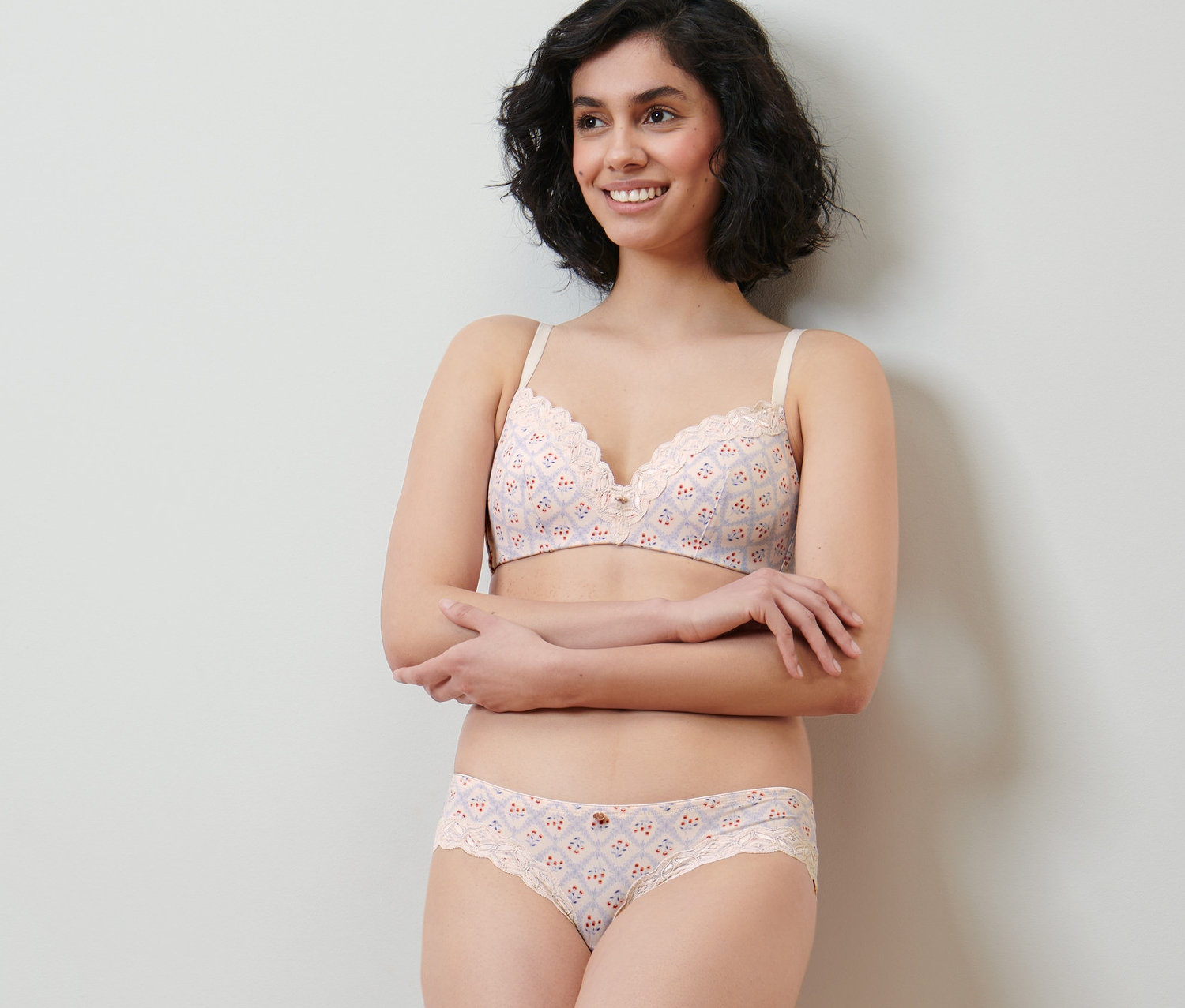 Usmívající se žena pózuje v krémové soupravě spodního prádla s potiskem a krajkou. Na sobě má brazilské kalhotky.