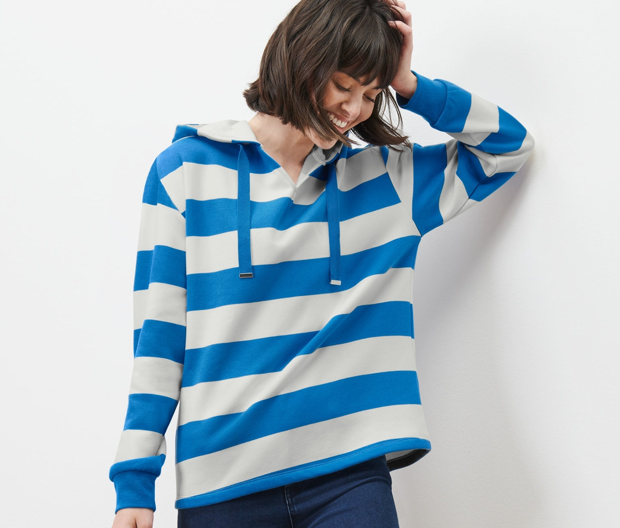 Žena opřená o zeď nosí pruhovanou mikinu s kapucí, džíny a tenisky.