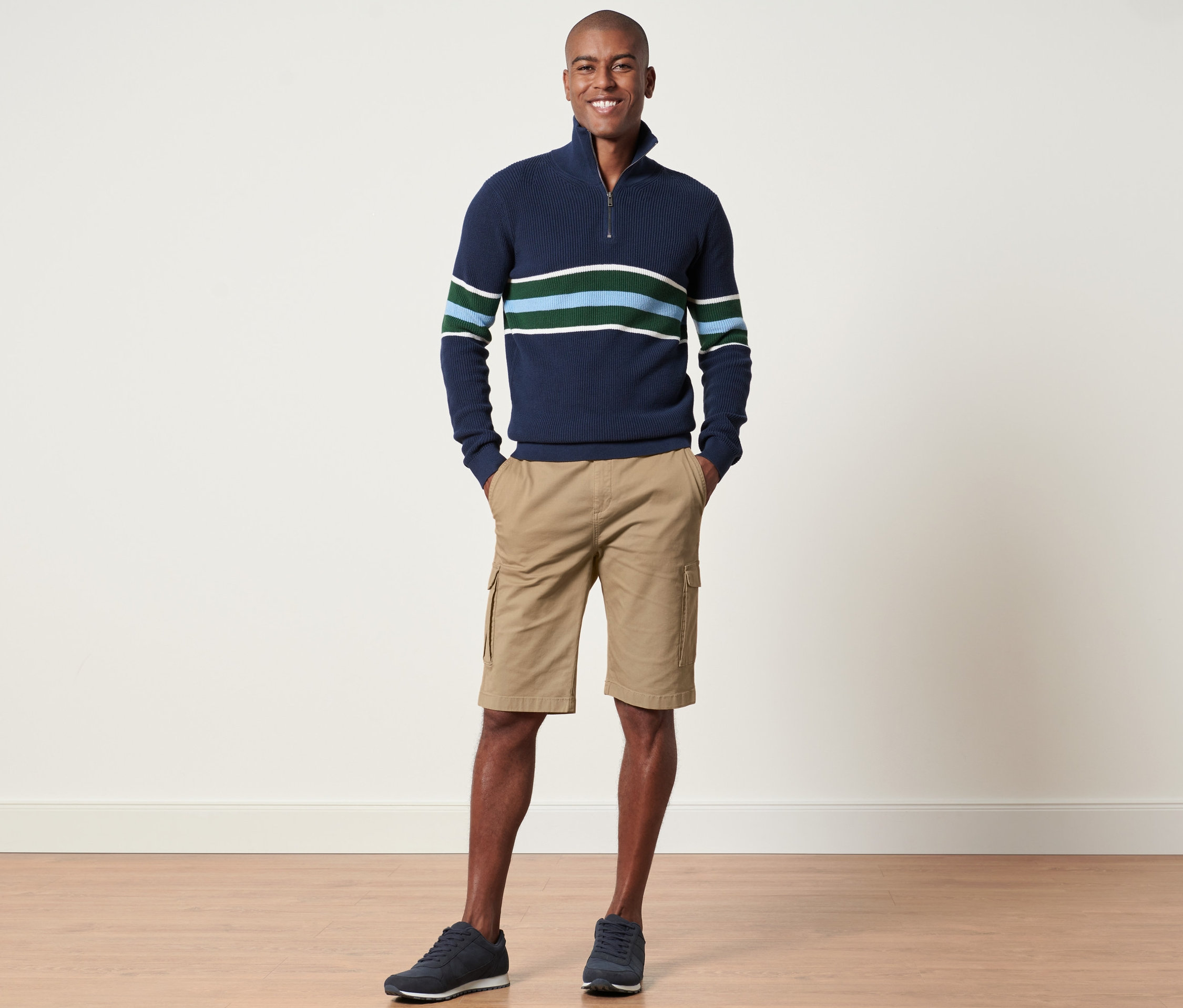 Muž stojí a pózuje v námořnickém svetru s pruhy a pískových cargo šortkách.