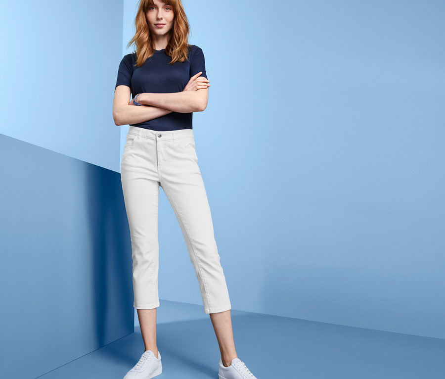 Žena pózuje v bílých džínách straightfit, modrém tričku a bílých botách na modrém pozadí.