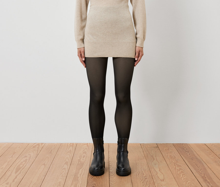 Žena má na sobě béžové šaty, černé punčochy a černé boty.