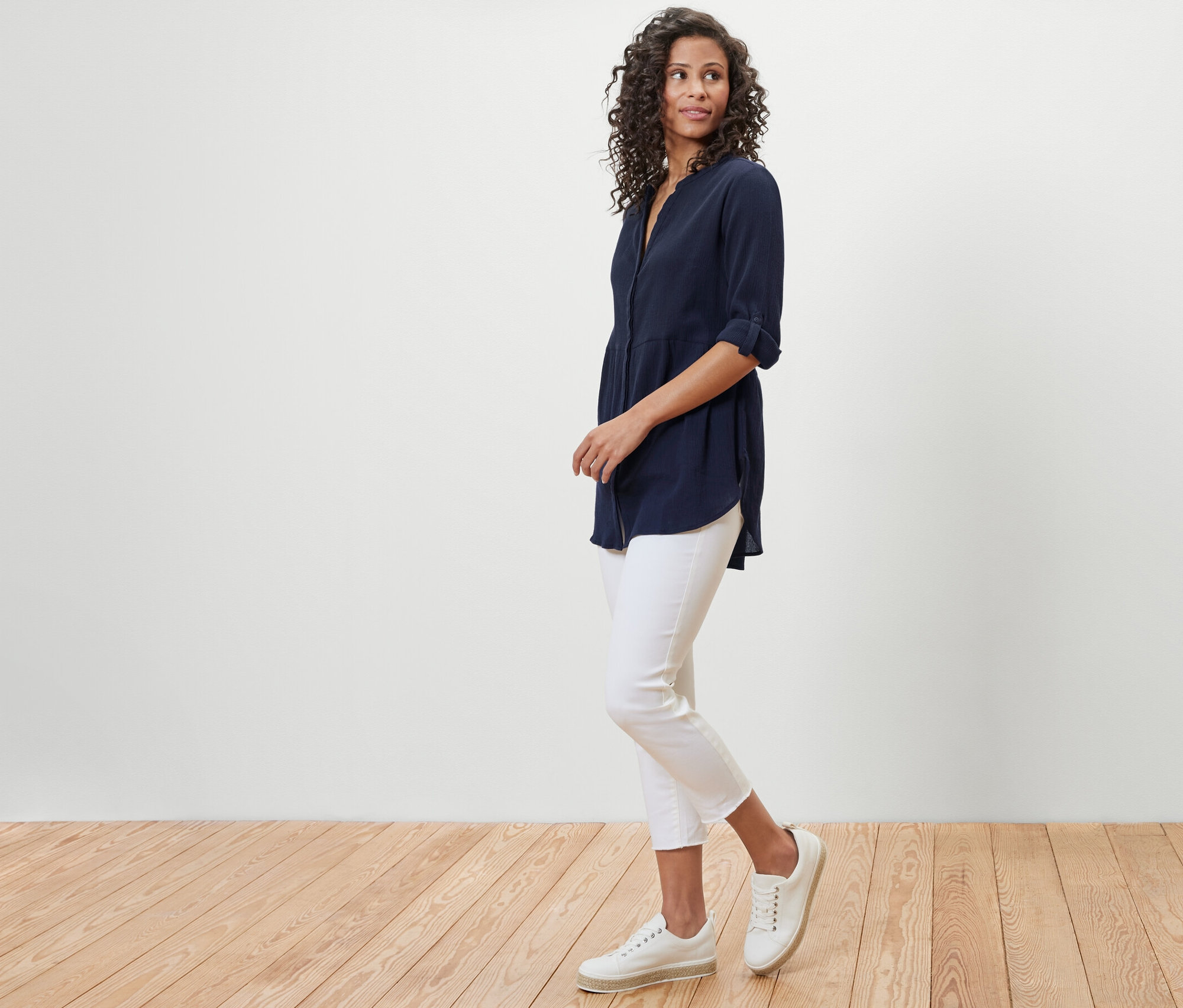 Žena v tmavě modré dlouhé halence a bílých kalhotách pózuje na dřevěné podlaze.