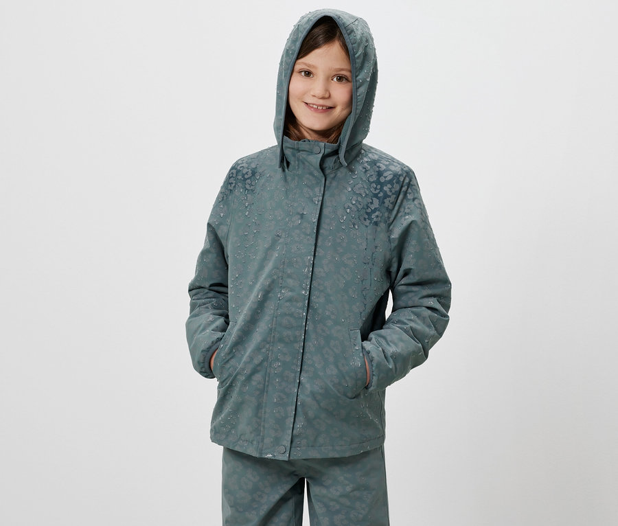 Usmívající se dívka pózuje v termo bundě do deště s kapucí a reflexními prvky.