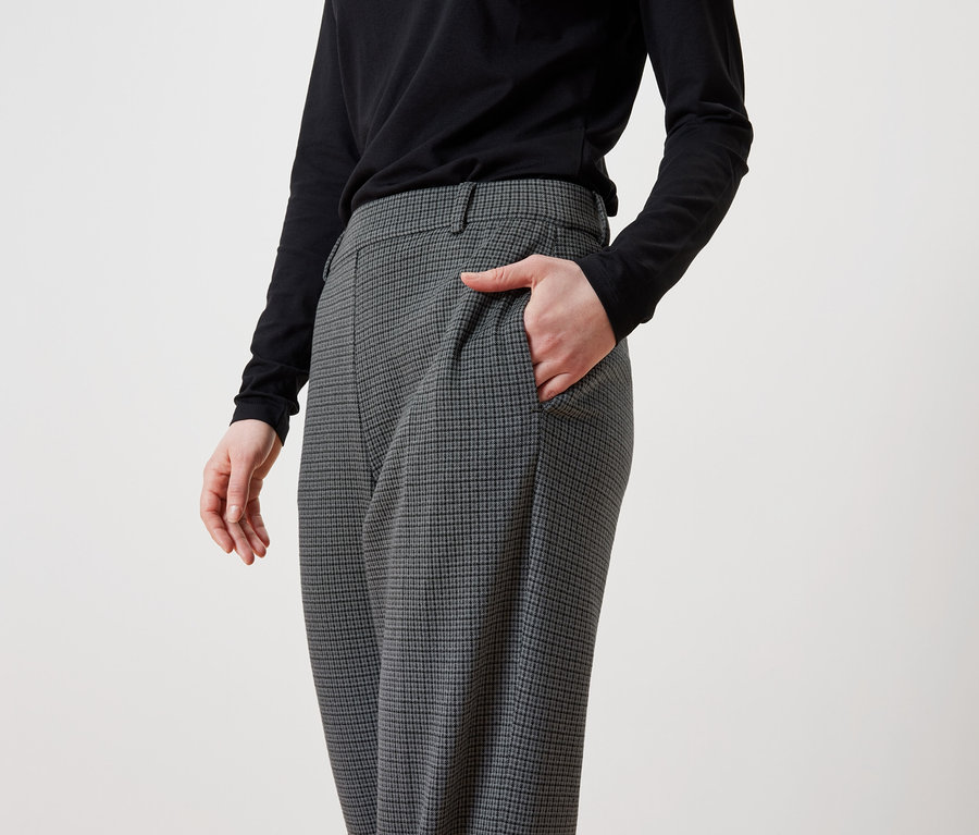 Žena má ruku v kapse šedých kostkovaných kalhot a má na sobě černé tričko s dlouhým rukávem.