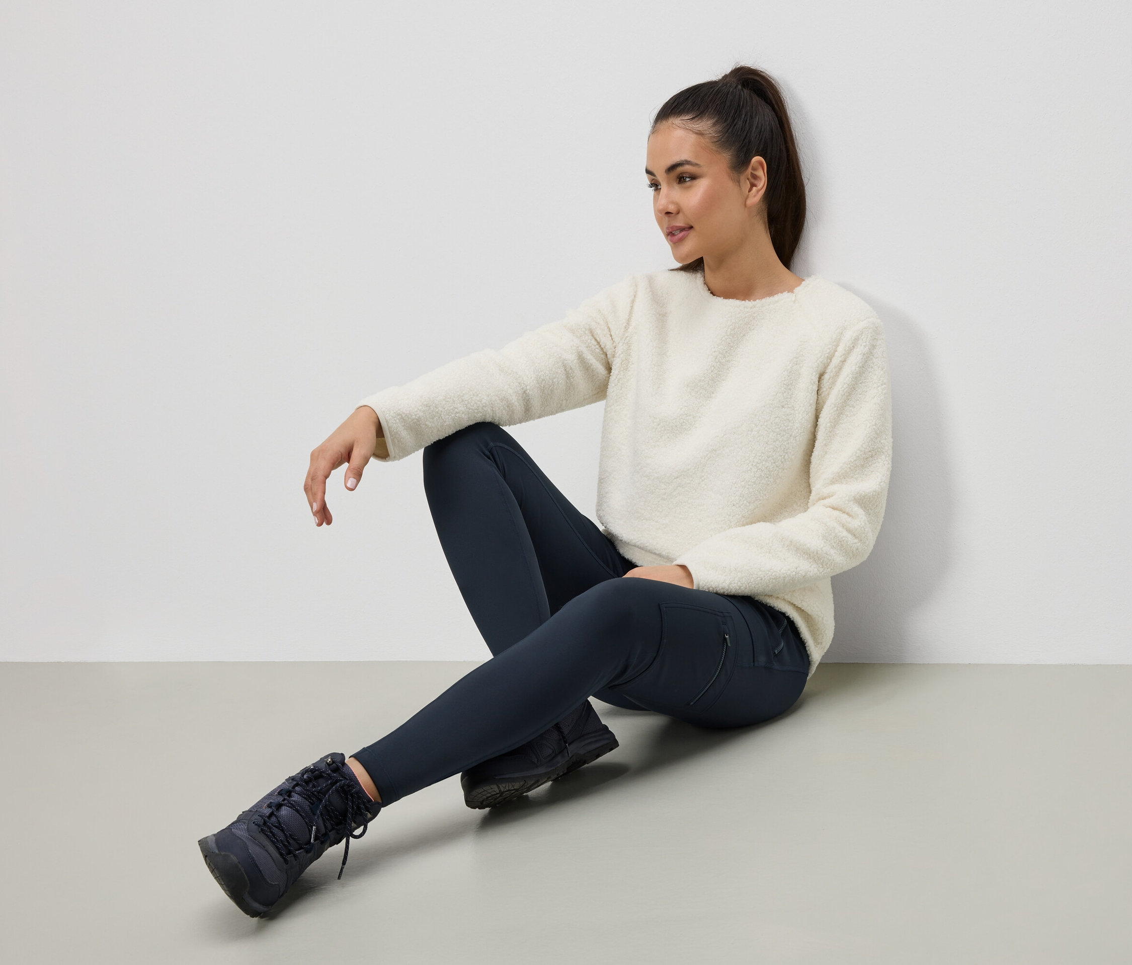 Žena sedí na podlaze v krémovém svetru, tmavě modrých legínách a botách.