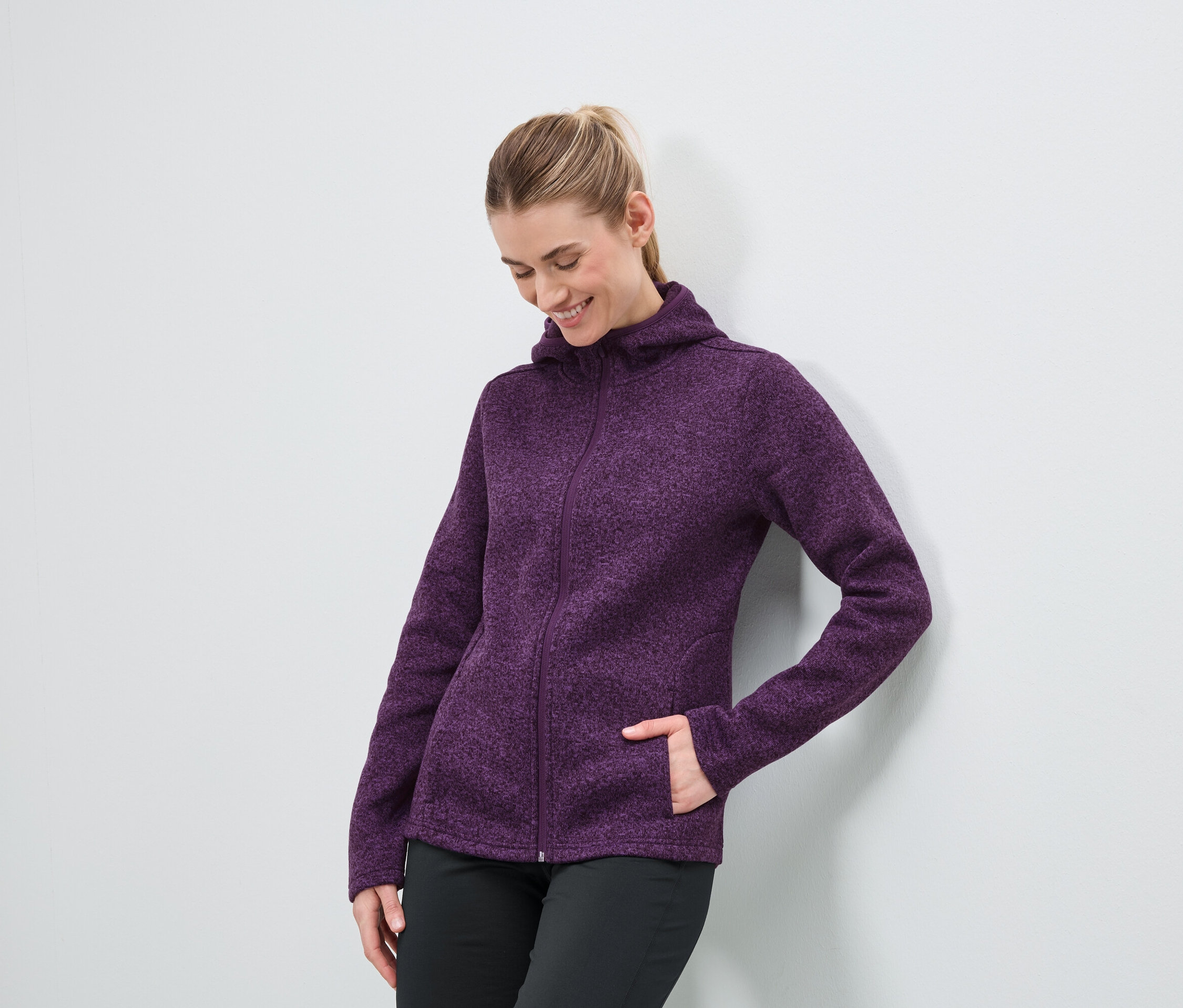 Žena v purpurové mikině s kapucí se zipem a černých kalhotách stojí u bílé zdi.
