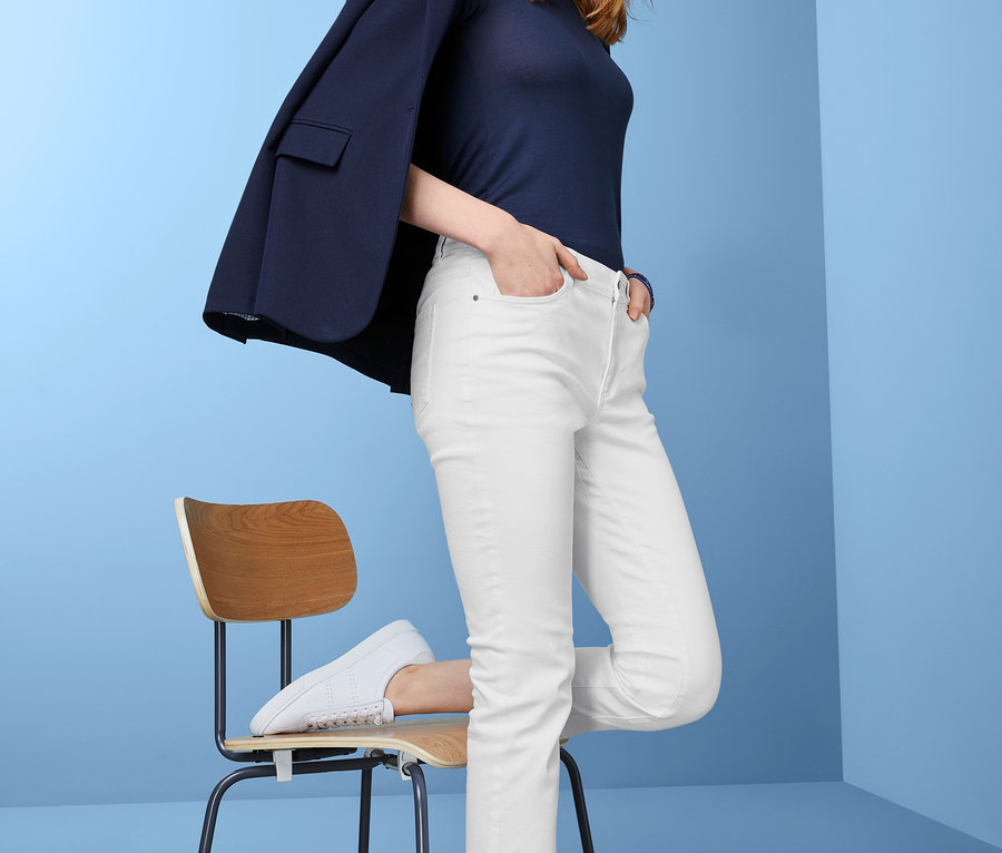 Žena pózuje v bílých džínách straightfit, tmavě modrém topu a saku přes rameno, opírajíc se o židli.