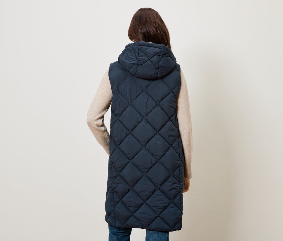 Žena zády k fotoaparátu má na sobě dlouhou prošívanou tmavě modrou vestu s kapucí.