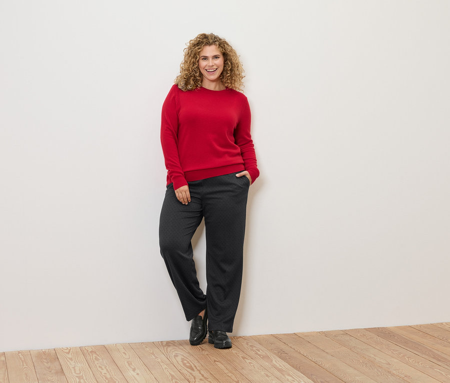 Žena s kudrnatými vlasy se opírá o zeď a má na sobě červený svetr a tmavé kalhoty.